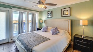 A bedroom with a large bed, a ceiling fan, a nightstand with a lamp, and a view of the outdoors through the window. - Walk to Beach! Pet-Friendly Mustang Island Retreat (Port Aransas)