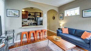 Ein Wohnzimmer mit Sofa, orangefarbenen Hocker, einem Couchtisch und einem durch einen Bogen sichtbaren Küchenbereich.