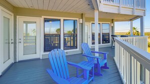 A covered porch with two blue Adirondack chairs, a white railing, and a view of the beach. - Beach Boardwalk Stay, 10-Min Walk, 2-Min Drive (Port Aransas)
