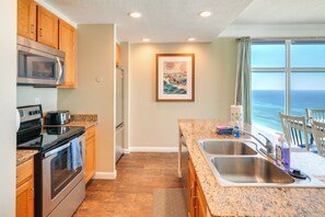 A kitchen with wooden cabinets, stainless steel appliances, a granite countertop, and a large window offering a view of the ocean. - Unwind Poolside, Walk to Shore! 12th-Floor PCB Gem (Panama City Beach)
