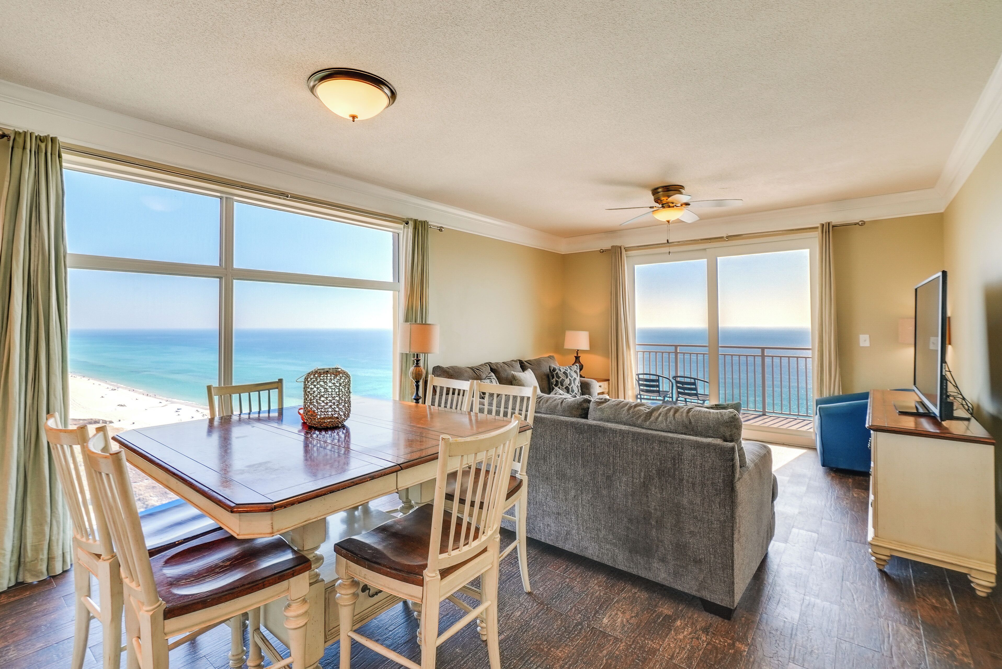 Quarto com mesa de jantar, cadeiras, sofá e televisão. Janelas amplas oferecem vista para a praia e oceano.