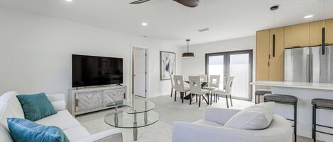 A modern living room with a flat-screen TV, white sofas, a glass coffee table, and a ceiling fan.
