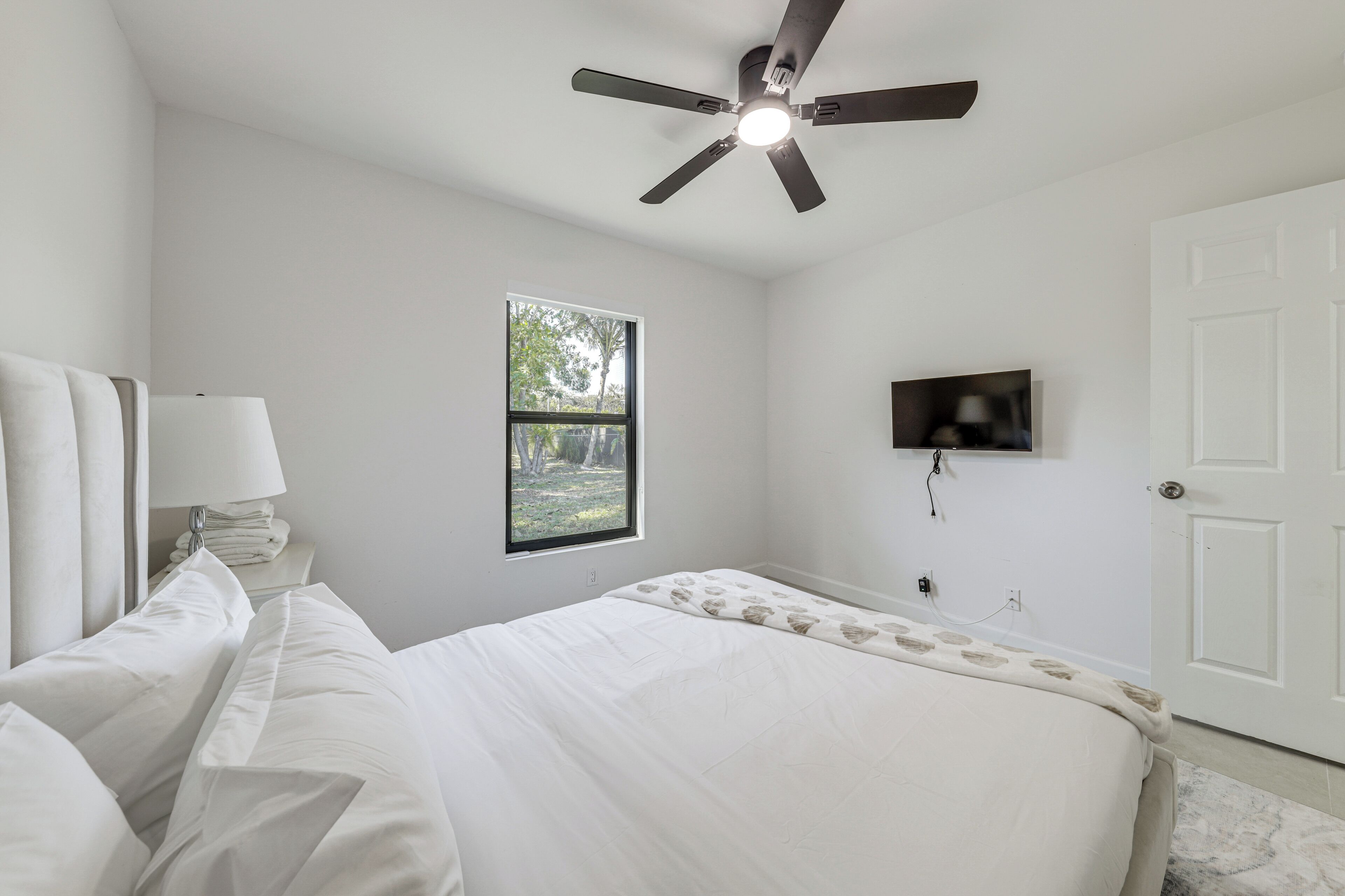 A bedroom with a bed, a ceiling fan, a window, a TV, and a door.