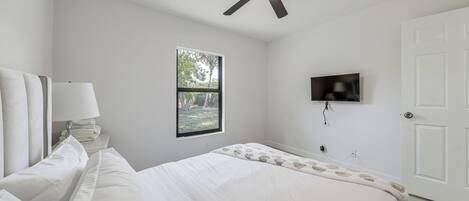 A bedroom with a bed, a ceiling fan, a window, a TV, and a door.
