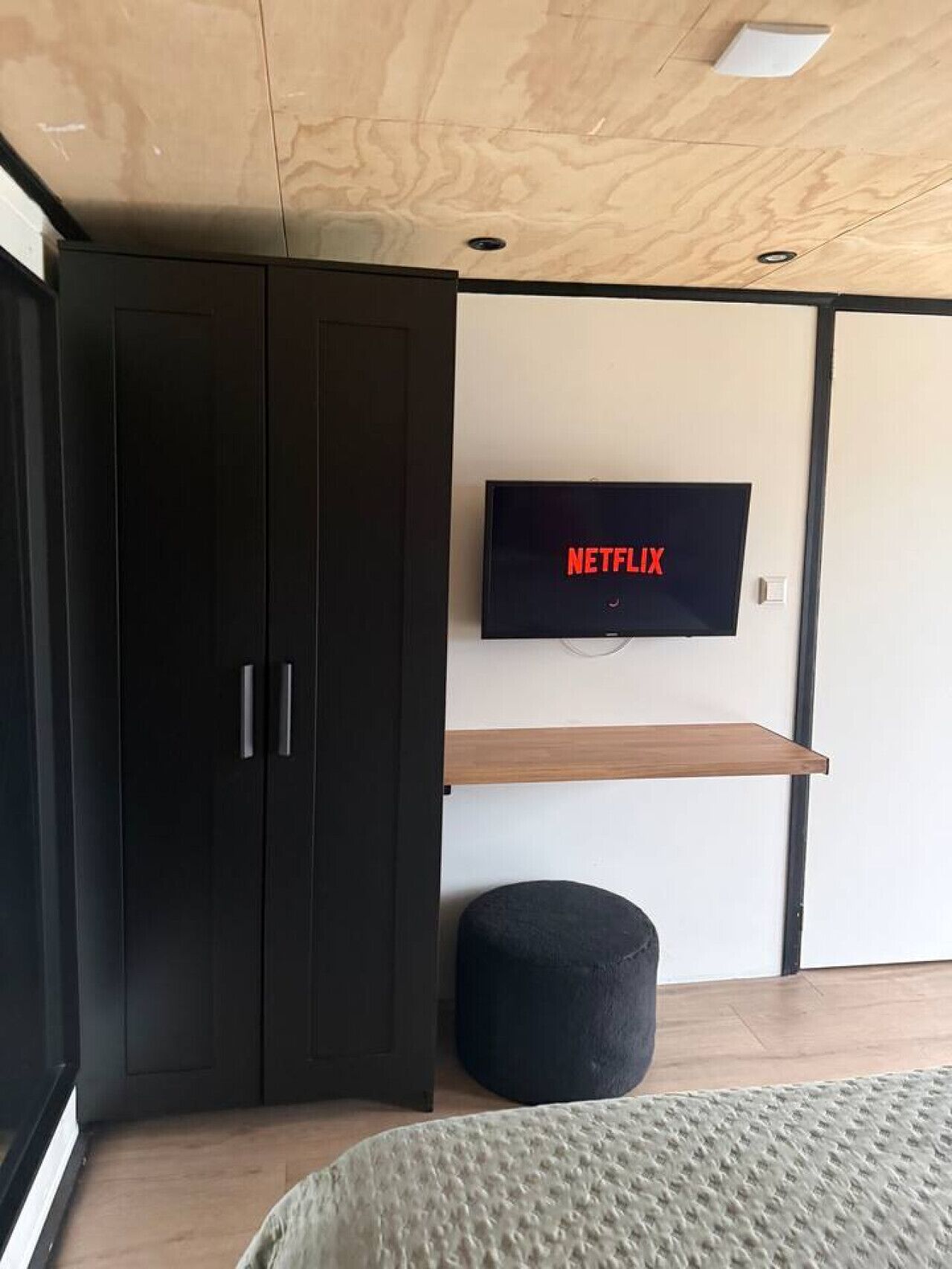 Una habitación con armario, televisor montado en la pared, estante de madera, otomana negra y cama con manta texturizada.