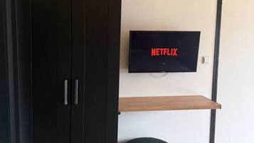 Una habitación con armario, televisor montado en la pared, estante de madera, otomana negra y cama con manta texturizada.