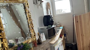Una cucina compatta con un piano di lavoro in marmo, un frigorifero bianco, un forno a microonde e un lavandino. Si nota uno specchio decorativo con cornice dorata e una ventola a parete.