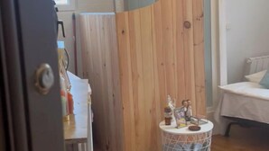 Una stanza con pavimento in legno, un piccolo tavolo rotondo con un cesto in filo di ferro e un letto con copriletto bianco.