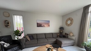 A modern living room with a grey sectional sofa, a black leather chair, a round coffee table, and a large window with blinds. - Holiday home for a family! (Katwijk aan Zee)