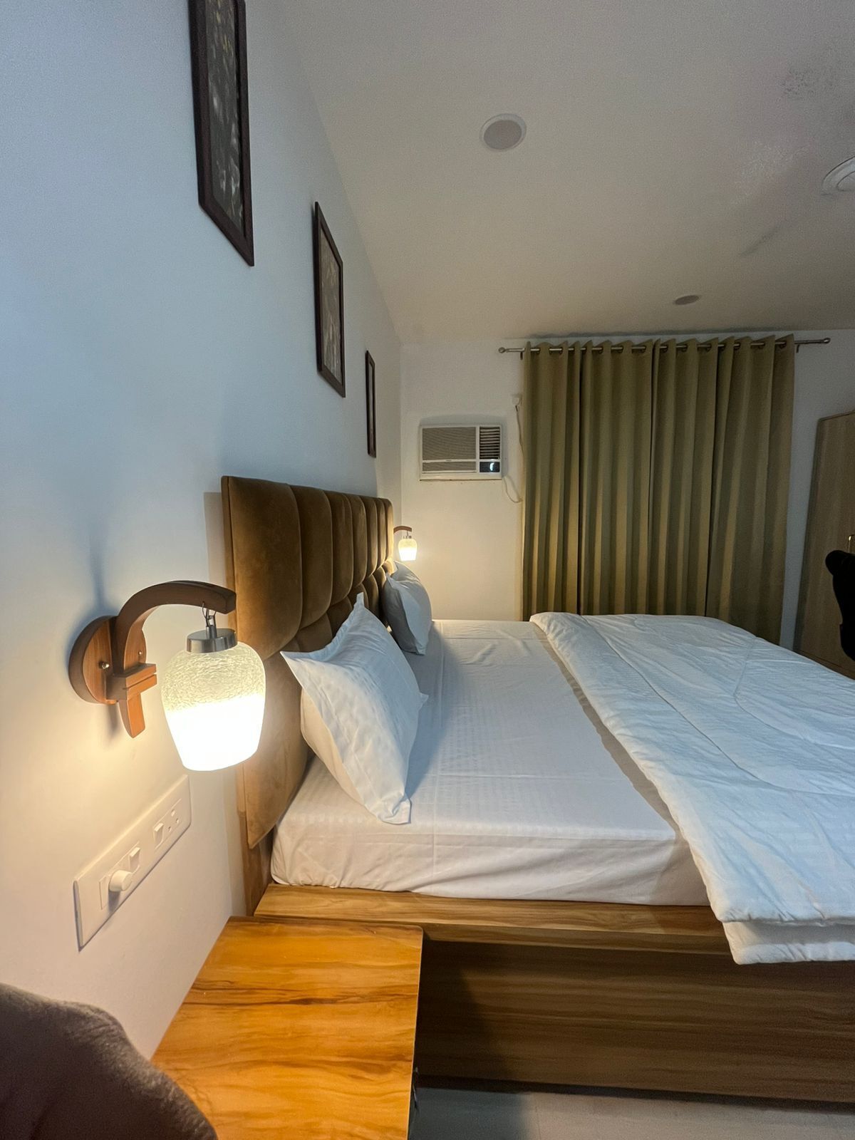 Une chambre d’hôtel comprenant un lit, un luminaire fixé au mur, une fenêtre avec des rideaux et un appareil de climatisation fixé au mur.