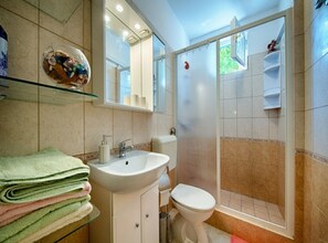 A bathroom with a glass shelf holding folded towels, a sink, a toilet, and a shower area with a glass door. - Golden Coast Apartments (Vis)