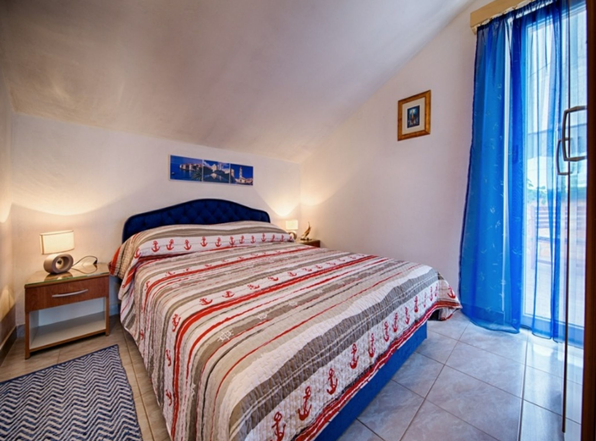 Quarto com cama, mesa de cabeceira, luminária, quadro na parede e janela com cortinas azuis.