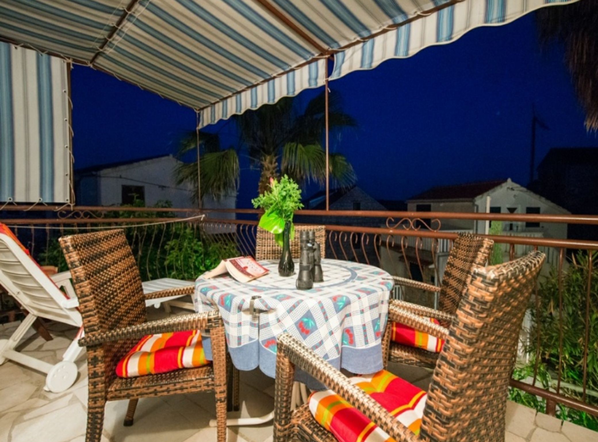 Een terras met rieten stoelen en een tafel, overdekt door een gestreepte luifel, met uitzicht op een woongebied.