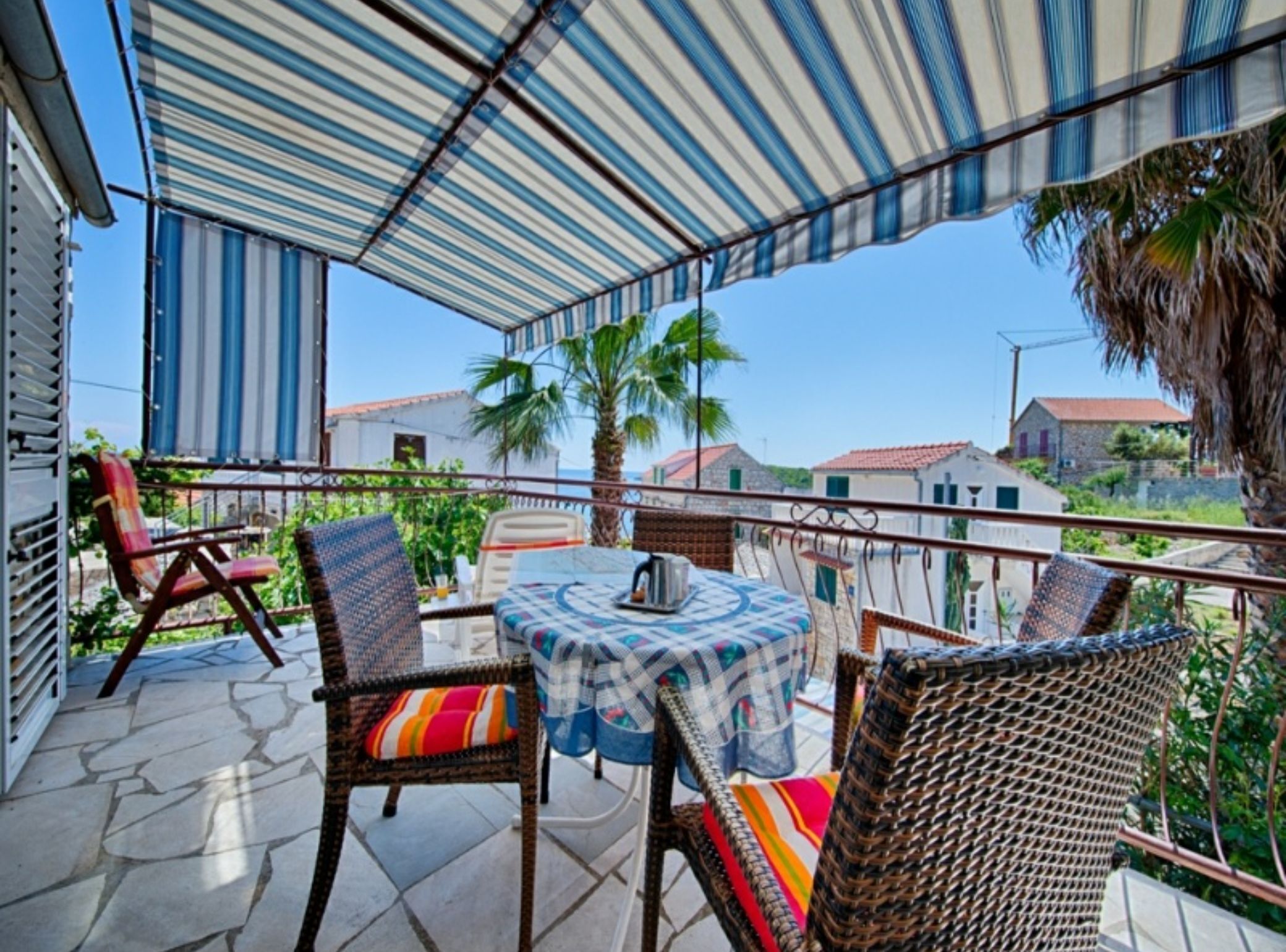 A covered outdoor seating area with wicker chairs and a table, overlooking a residential neighborhood with palm trees and houses.