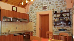 Una cocina con armarios de madera, una estufa y una mesa de comedor con sillas. Se aprecia una pared de piedra y una puerta al fondo.