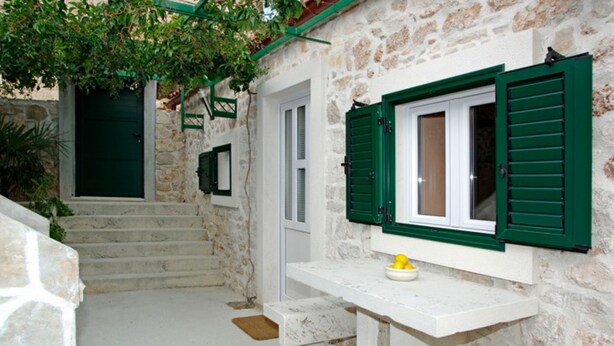 Un edificio de paredes de piedra con contraventanas verdes, una puerta blanca y una pequeña zona de estar al aire libre con un cuenco de limones.