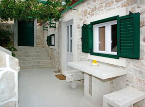 Un bâtiment aux murs de pierre, avec des volets verts, une porte blanche et un petit espace extérieur aménagé avec quelques chaises, où l’on peut voir un bol rempli de citrons.