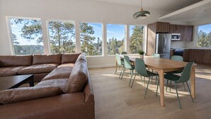 Ein modernes Wohnzimmer mit einer braunen Ledersofa, einem Holzesstisch mit Stühlen und großen Fenstern mit Blick auf Bäume und Hügel.
