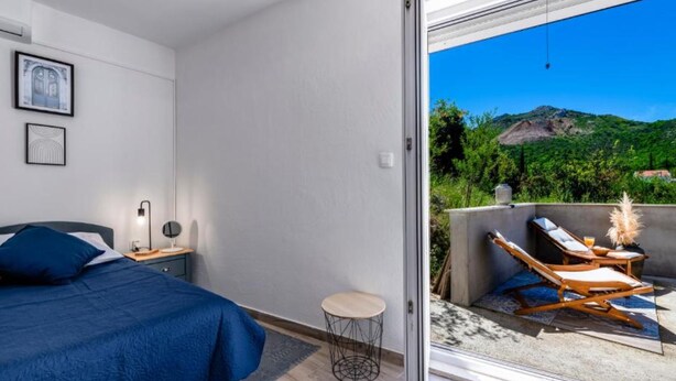 A bedroom with a bed, bedside table, lamp, and artwork. It features a sliding glass door leading to an outdoor seating area with a view of a mountainous landscape. - Lux Holiday Home Maribu (Zupa dubrovacka)