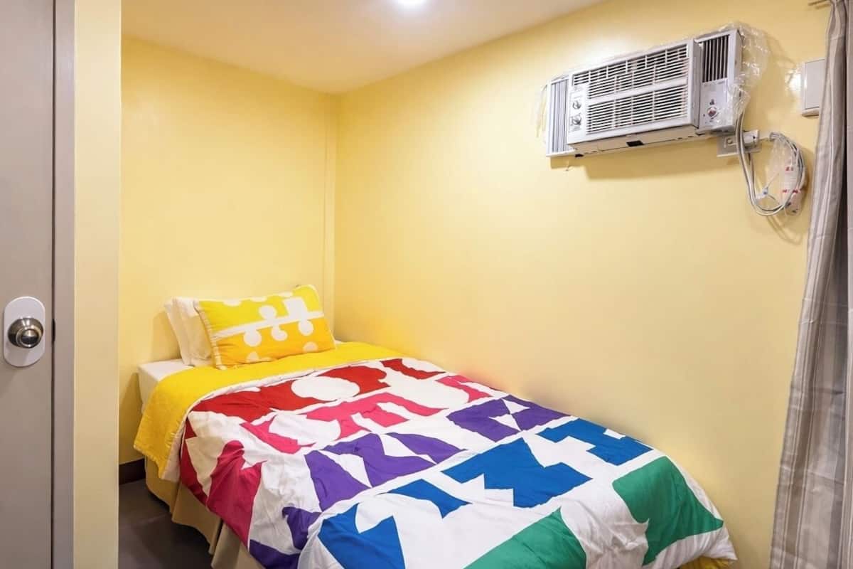 A small, single-bed room with a yellow wall, a colorful quilt, and a wall-mounted air conditioning unit.