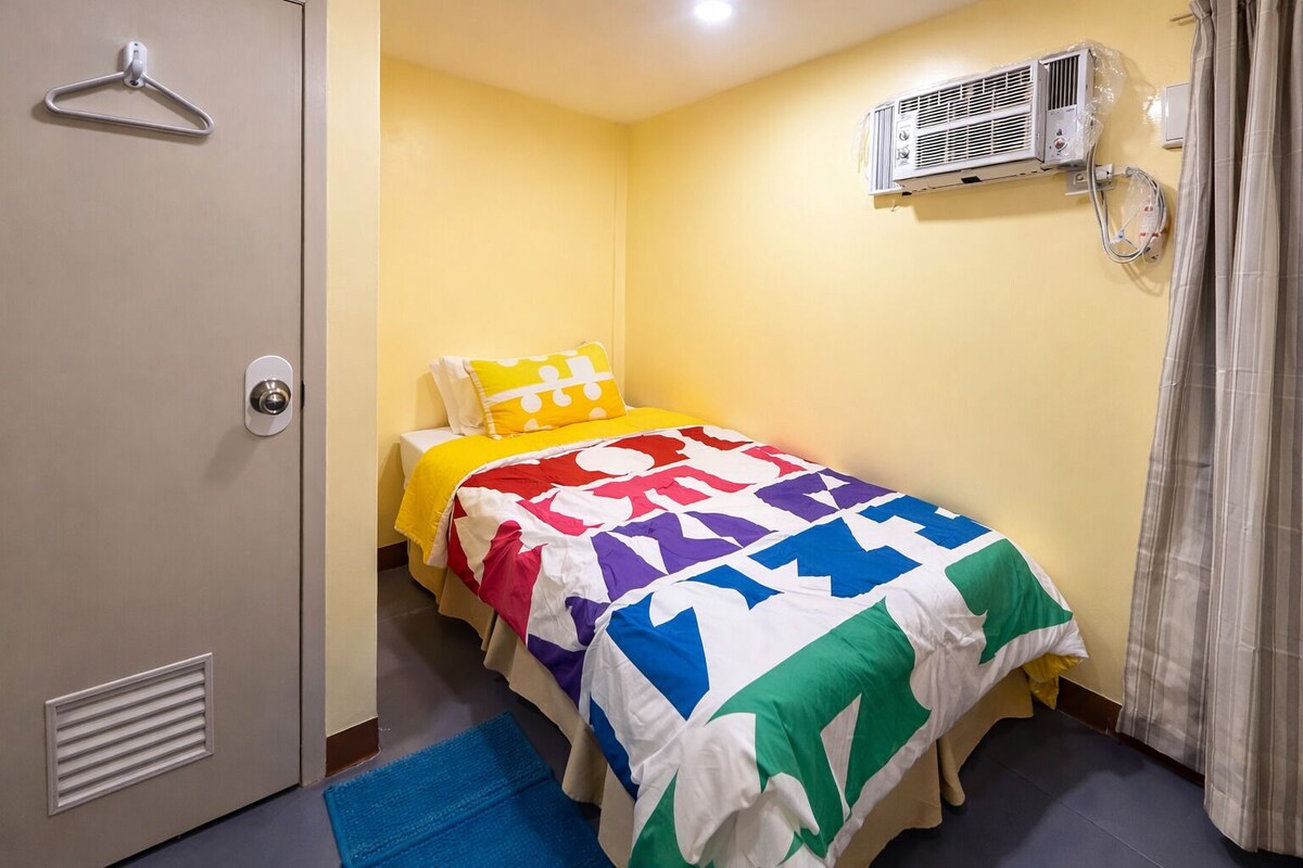 A small, single-bed room with a yellow wall, a bed with a colorful quilt, a white air conditioning unit, and a door with a hanger.