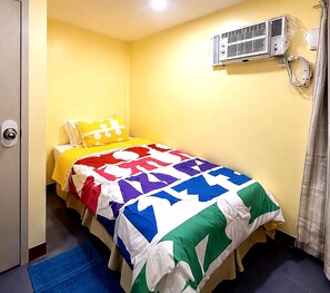A small, single-bed room with a yellow wall, a bed with a colorful quilt, a bedside air conditioning unit, and a blue mat on the floor.