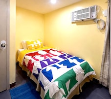 Una habitación pequeña con cama individual, pared amarilla, cama con colcha colorida, unidad de aire acondicionado junto a la cama y un tapete azul en el suelo.