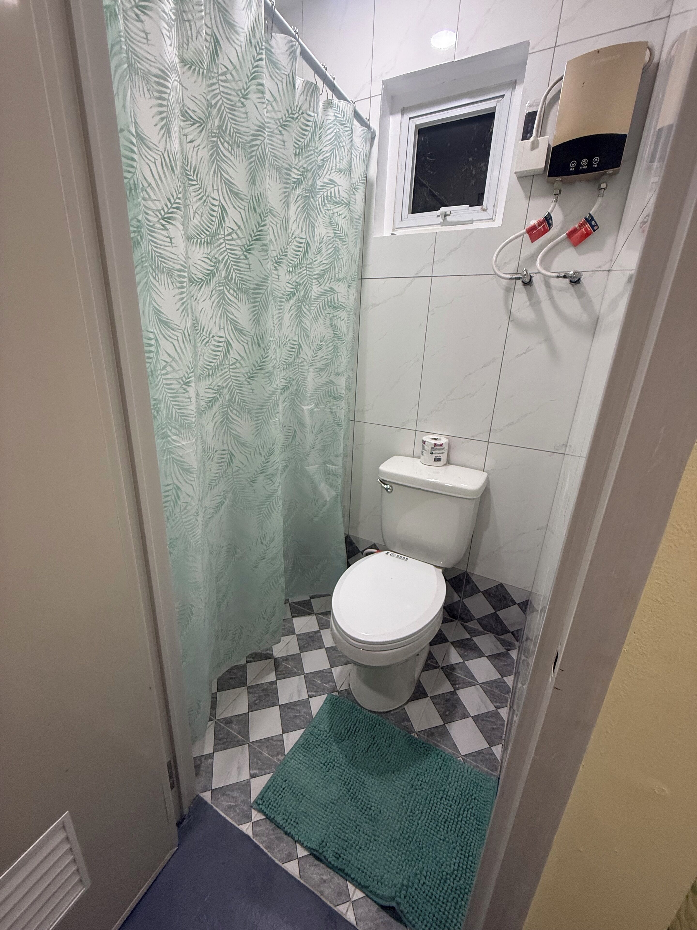 A small bathroom with a toilet, a shower curtain, a window, and a wall-mounted heater.