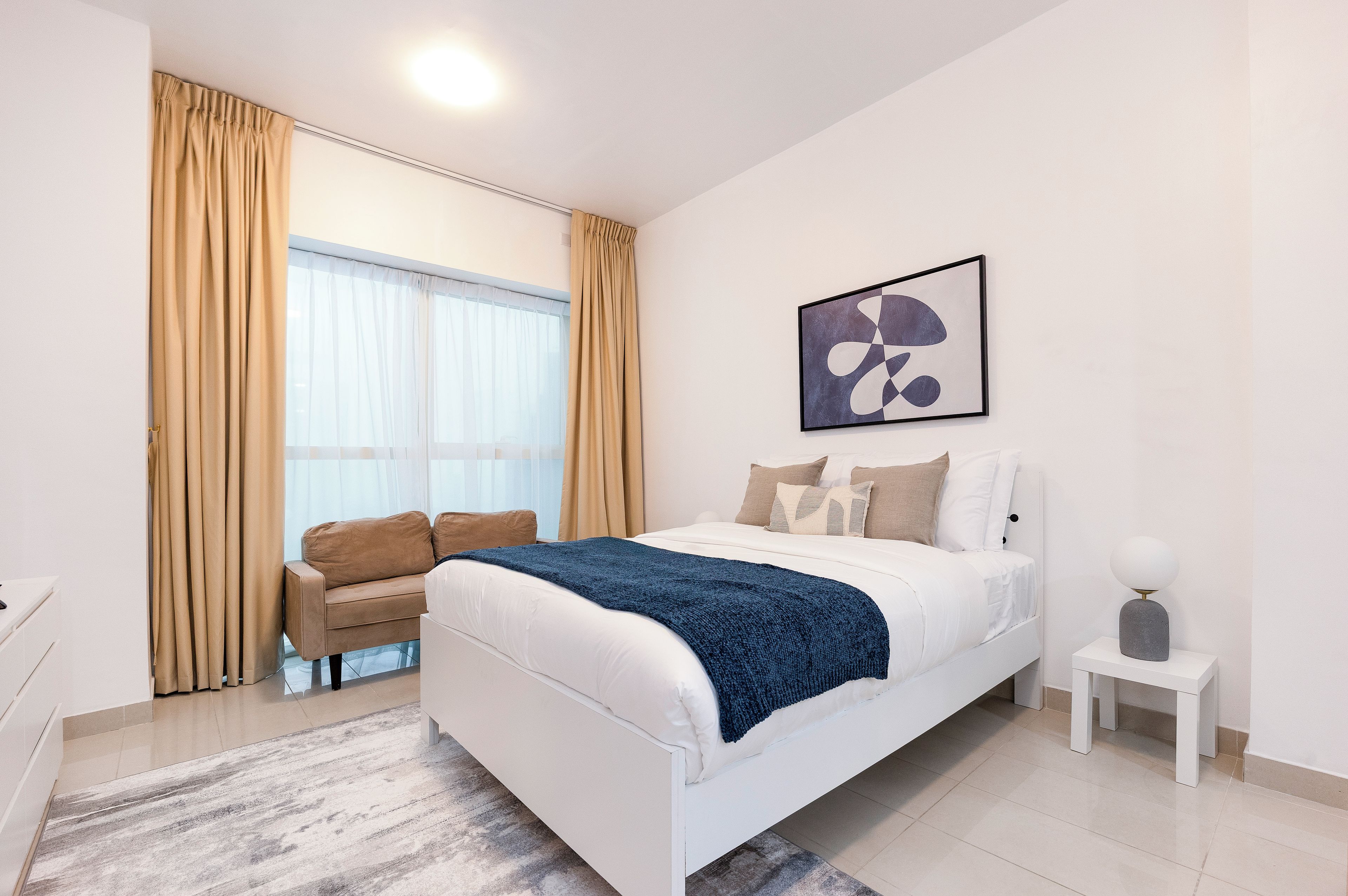 A modern bedroom with a bed, a sofa, a bedside table, and a large window with curtains.