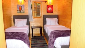Quarto de cabana de madeira com duas camas, uma mesinha e uma janela.