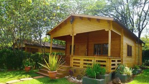 Uma cabana de madeira com varanda, cercada por um jardim bem cuidado com diversas plantas e um caminho de pedra que leva à entrada.