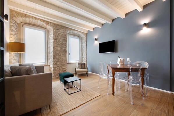 A modern living room with a stone wall, wooden ceiling beams, a flat-screen TV, a dining table with clear chairs, and a sofa. - Il Vicolo Suite Apartments Cortiletto (Gardola)