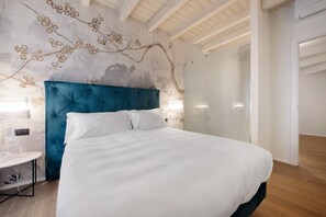 A bedroom with a large bed, a blue upholstered headboard, a wall with floral wallpaper, and a wooden floor. - Il Vicolo Suite Apartments Cortiletto (Gardola)