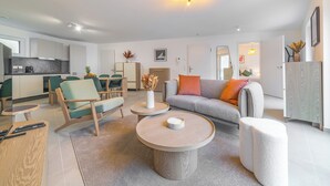 A modern living room with a grey sofa, teal armchairs, and a round coffee table. A kitchen with white cabinets and a dining area with a table and chairs are visible in the background. - Halternative Residence Rose (Luxembourg City)