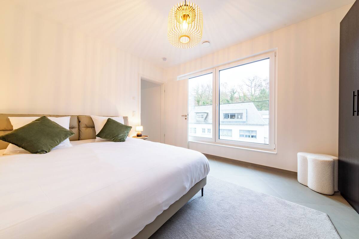 Ein modernes Schlafzimmer mit einem großen Bett, einem Nachttisch, einer Lampe und Blick ins Freie durch ein großes Fenster.