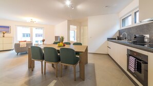 A modern kitchen with a dining area, featuring a wooden table and chairs, a built-in oven, and a large window providing natural light. - Halternative Residence Rose (Luxembourg City)
