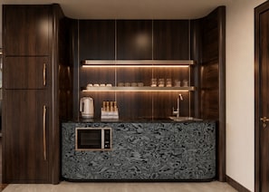 A modern kitchen with dark wood cabinetry, a marble-patterned countertop, a built-in microwave, and a shelf with glassware and a coffee maker. - The Prajan Hotel & Villas by Natt's (Yogyakarta)