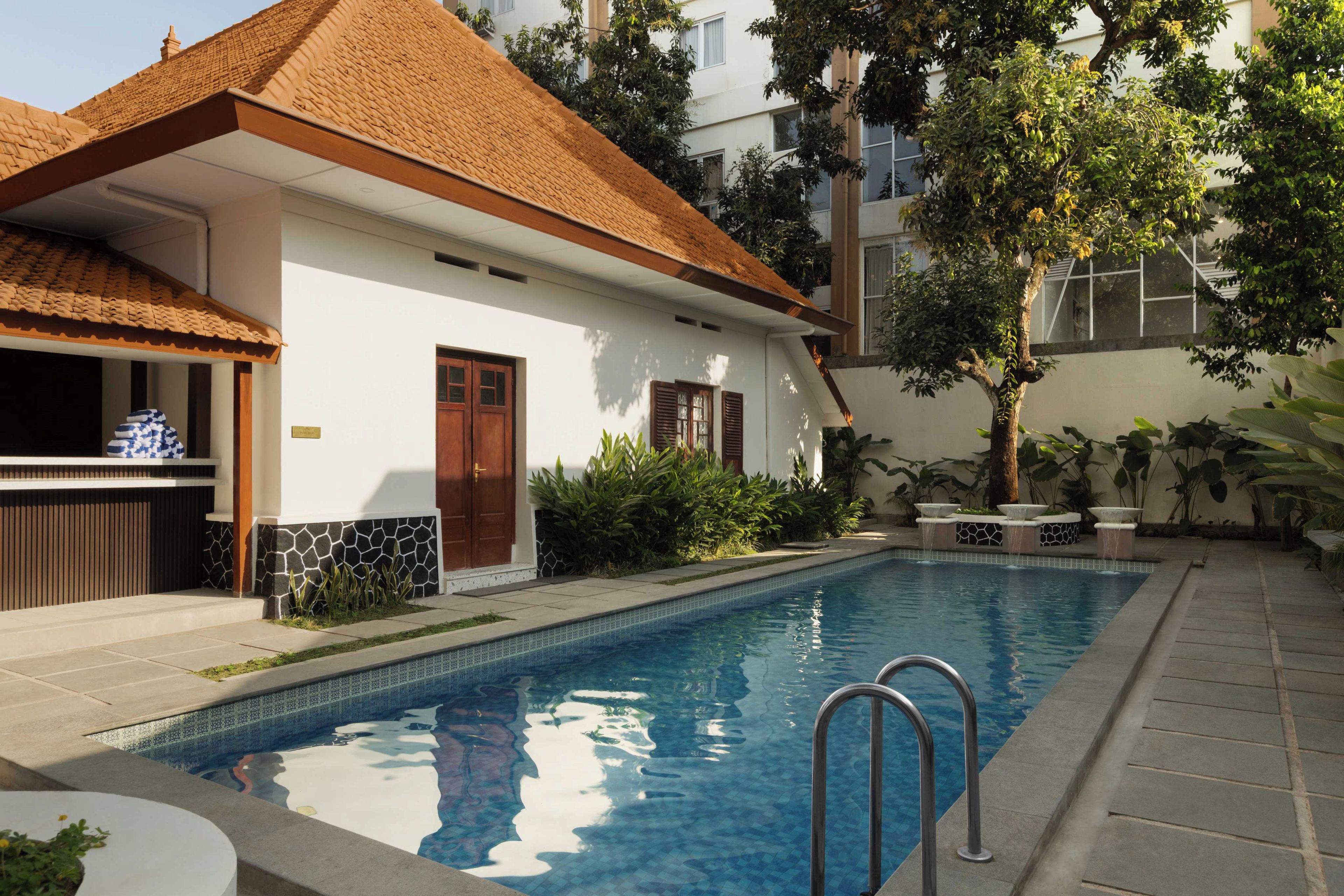 Una piscina con borde de azulejos, una puerta de madera y un edificio blanco con techo de tejas.