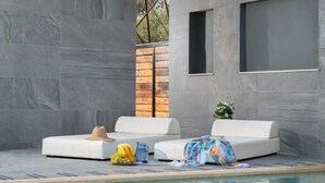 Ein moderner Poolbereich im Freien mit zwei weißen Liegestühlen, einem Holzzaun und einem kleinen Fenster.