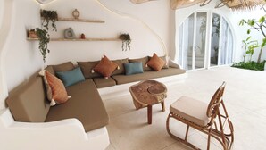 A modern living room with a sectional sofa, a wooden coffee table, and a wicker chair. There are plants on shelves and a large window with a view. - Suilaya Villa Uluwatu (Pecatu)