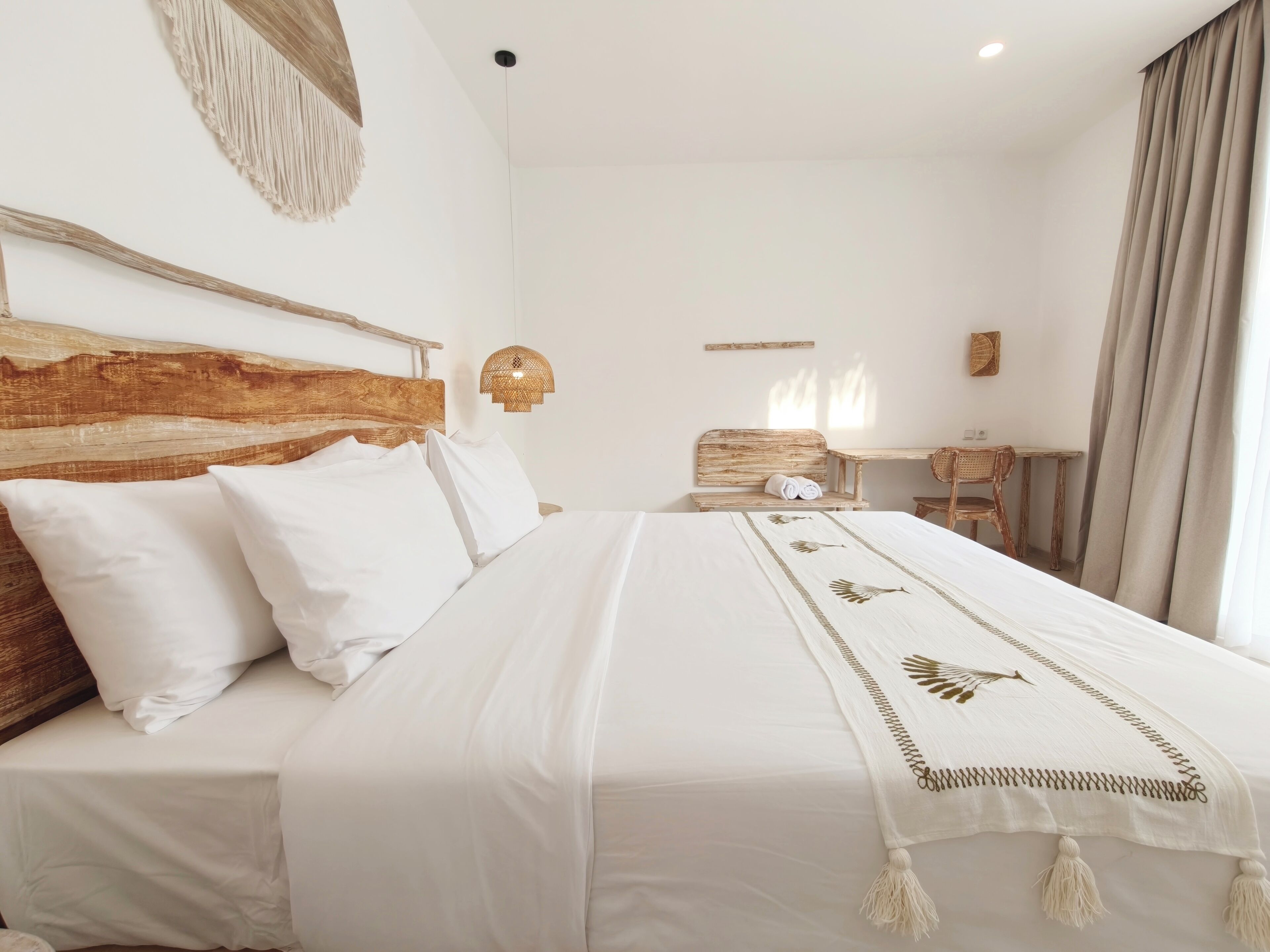 A neatly made bed with white linens and a decorative throw, a wooden headboard, and a wicker chair in a bright room with a simple, clean design.