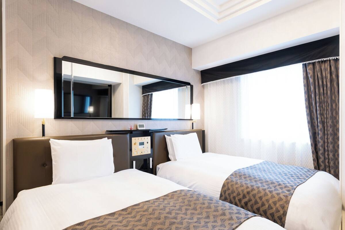 Ein Hotelzimmer mit zwei Betten, einem großen Spiegel, einer Bedienkonsole und einem Fenster mit Vorhängen.