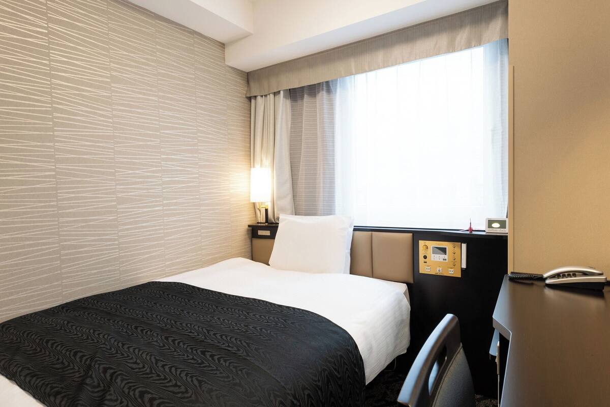Ein Hotelzimmer mit Bett, Nachttisch mit Lampe, Bedienfeld und einem Fenster mit Vorhängen.