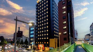 Un hotel moderno con un ingresso ben visibile, un ponte pedonale verde e una trafficata via cittadina al crepuscolo.