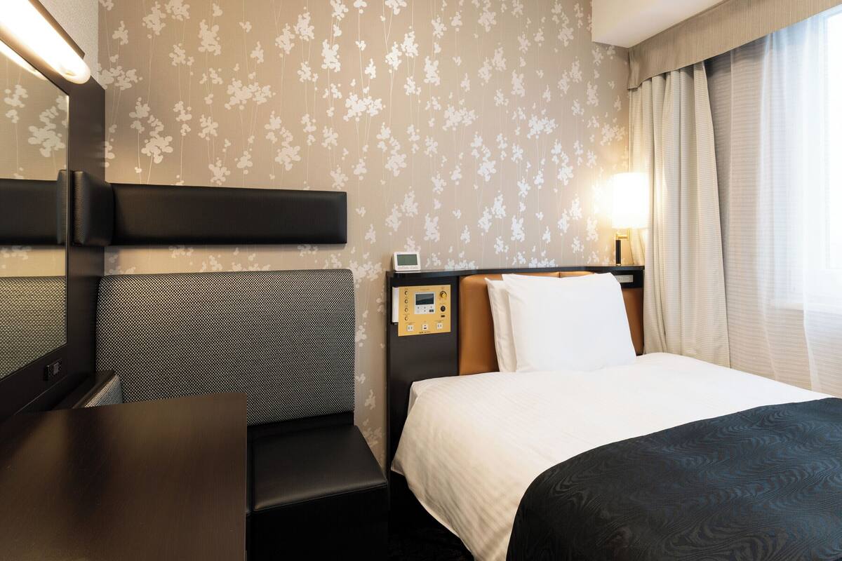 Ein Hotelzimmer mit Bett, Schreibtisch, Kopfteil, Nachttisch, Lampe und einem Fenster mit Vorhängen.