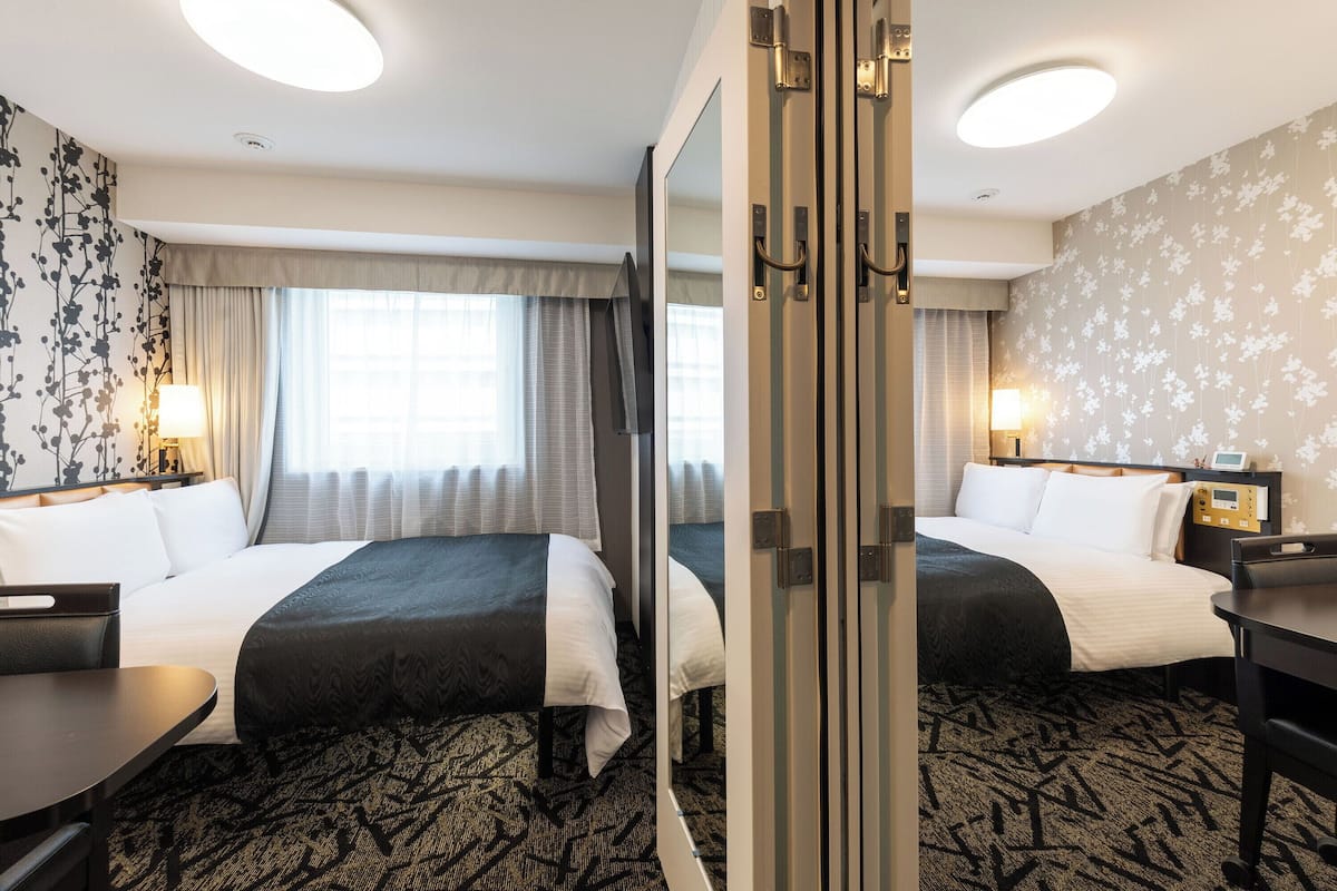A hotel room with two beds, a desk, a mirror, and a nightstand with a lamp.