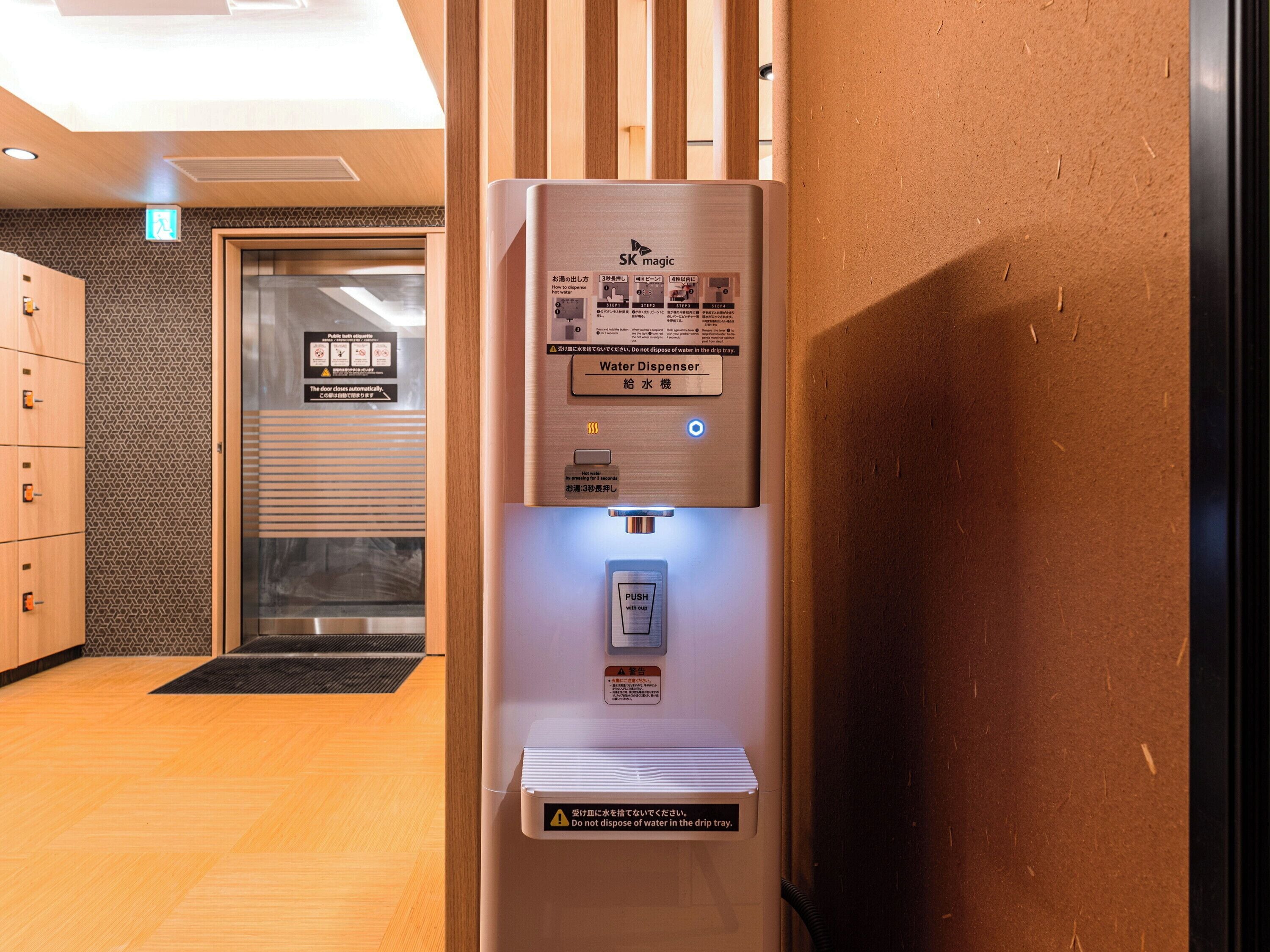 A water dispenser with instructions in multiple languages, a push button for hot water, and a warning not to dispose of water in the drip tray.