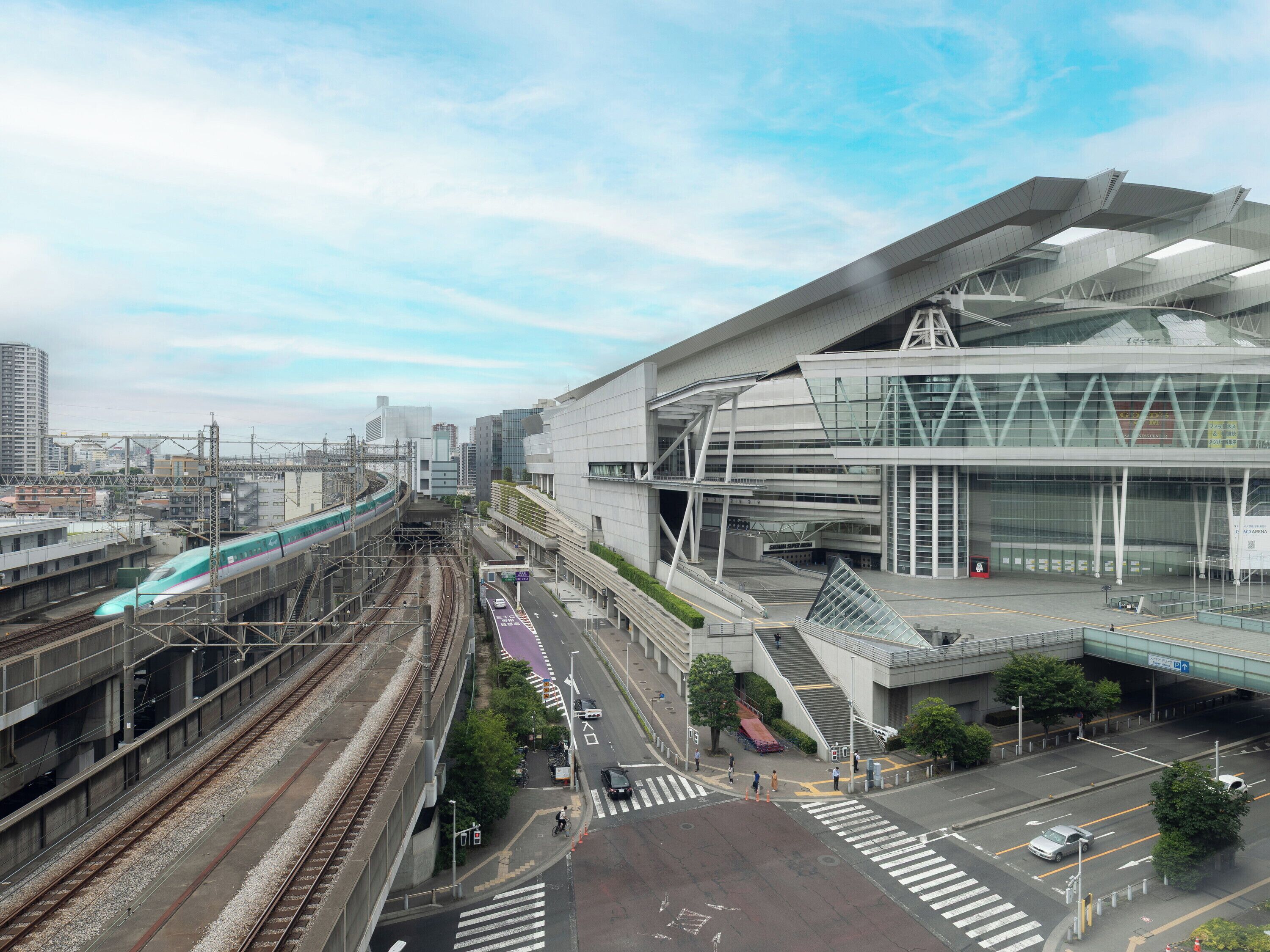 A modern train station with multiple tracks, a pedestrian bridge, and a large building with a unique architectural design.