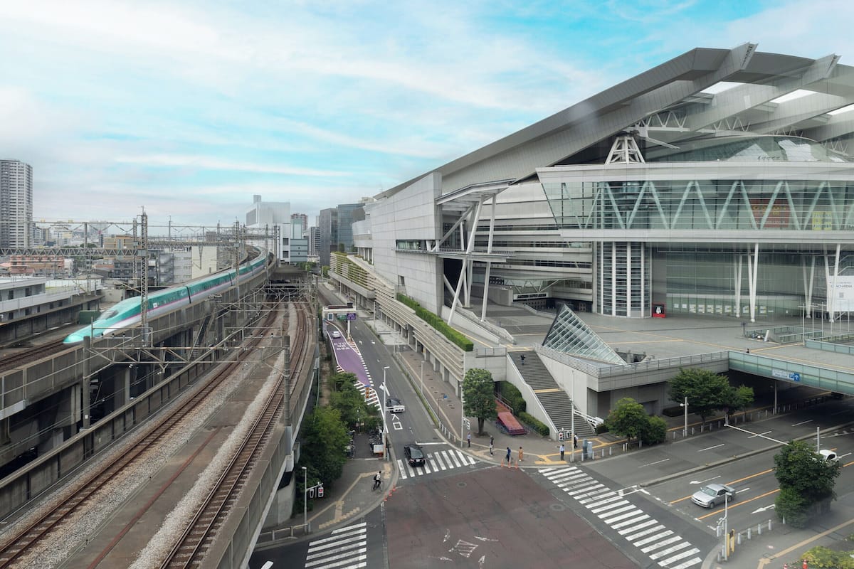 A modern train station with multiple tracks, a pedestrian bridge, and a large building with a unique architectural design.