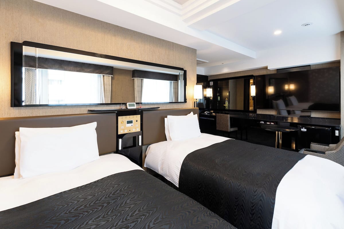 A hotel room with two beds, a desk, a chair, a mirror, and a control panel.